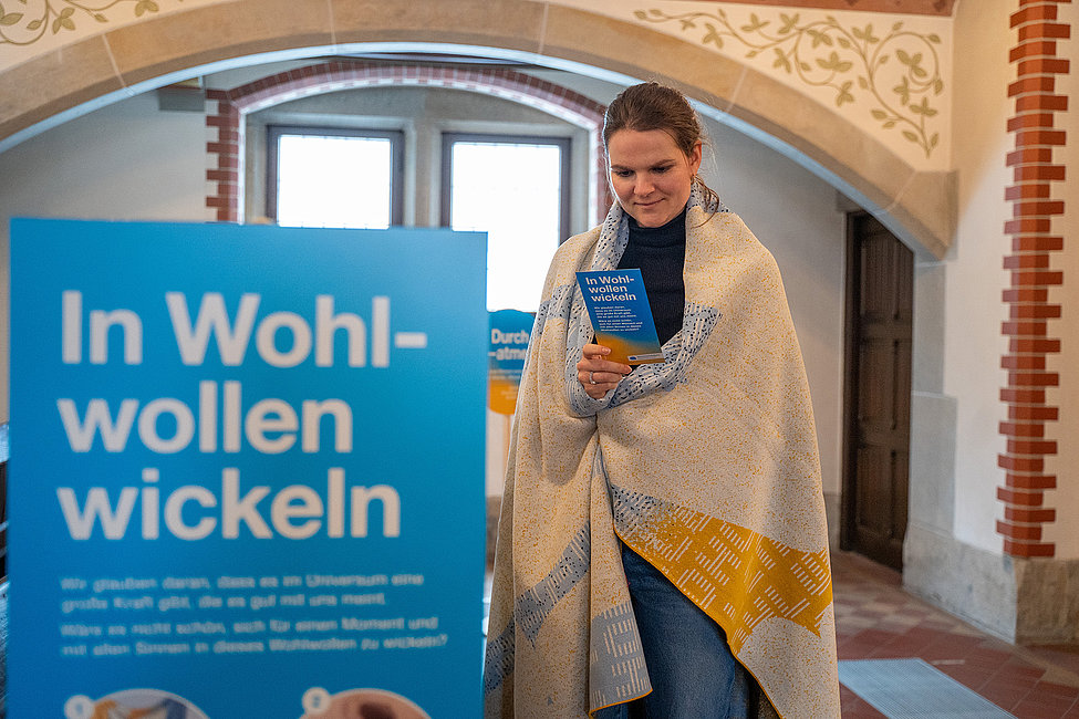 Frau steht eingewickelt in eine Decke und liest einen Flyer mit der Aufschrift „In Wohlwollen wickeln“ in einem historischen Gebäude.