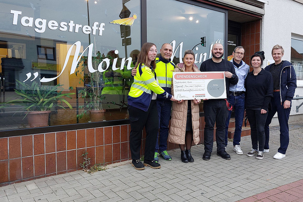 Eine Gruppe von Menschen steht vor einem Gebäude mit der Aufschrift "Tagestreff Moin Moin" und hält gemeinsam einen großen Spendenscheck. Alle lächeln in die Kamera. Die Szene wirkt freundlich und unterstützend.