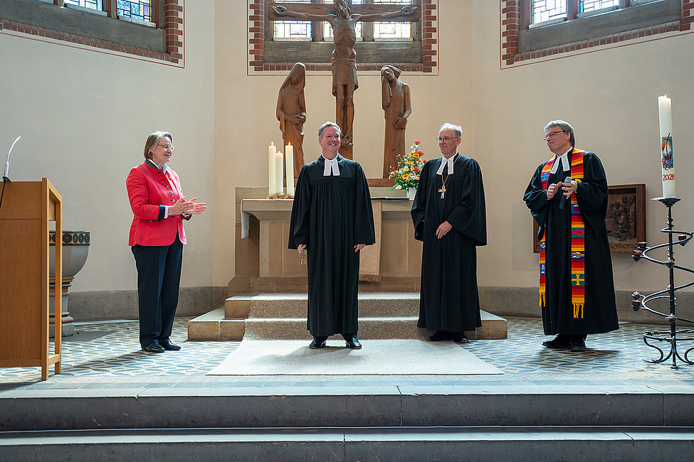 Vier Personen in liturgischer Kleidung stehen am Altar einer Kirche; eine Frau rechts applaudiert, Kreuz und Kerzen im Hintergrund.