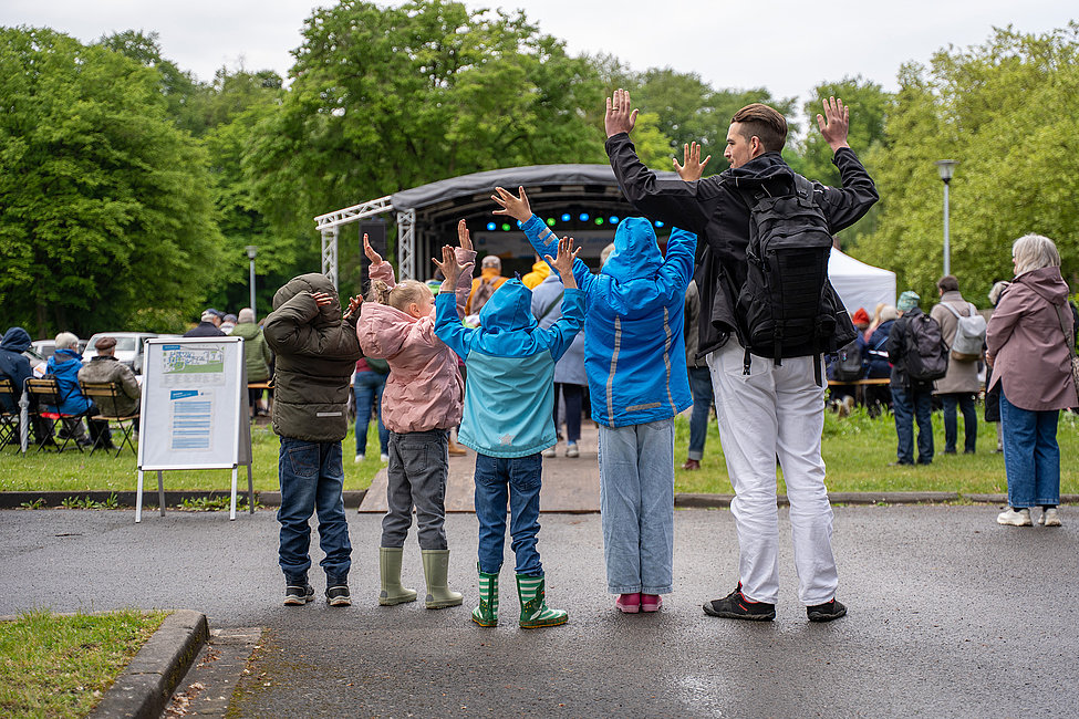 Zu sehen sind vier Kinder und ein Mann von hinten, alle tragen Regenkleidung und heben die Arme nach oben. Im Hintergrund ist eine Bühne zu erkennen.