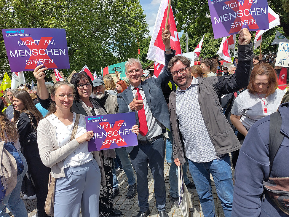 Menschen demonstrieren im Freien; mehrere halten Schilder „Nicht am Menschen sparen“, einige lächeln in die Kamera, bunte Fahnen im Hintergrund.
