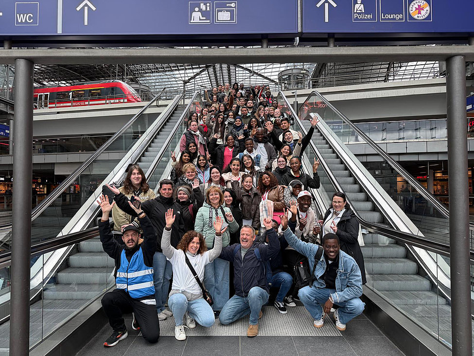 Viele Menschen sitzen auf einer Treppe am Bahnhof, schauen fröhlich und heben ihre Hände hoch.